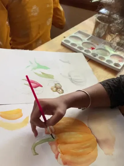 A close-up of a student's hand as they paint a pumpkin. This shot highlights the delicate brushwork and layering involved in watercolor still life.