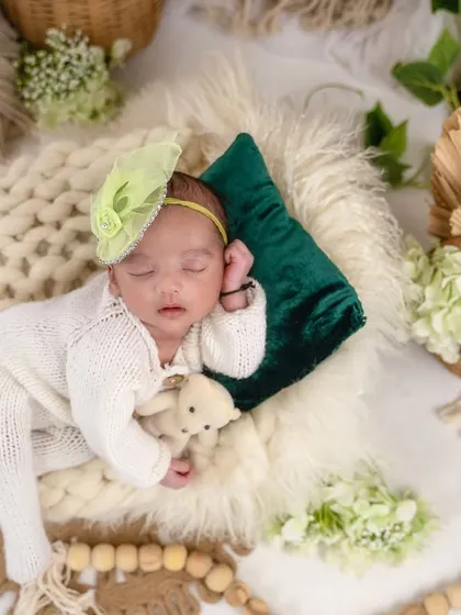 A beautiful boho setup featuring a baby in a white knit outfit sleeping on a chunky wool blanket. The scene is decorated with wicker baskets, dried palm leaves, and green foliage.