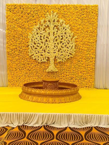 A simple and classic Haldi stage. The backdrop is made of a wall of yellow marigolds with a 'Tree of Life' motif created from white flowers, providing a bright and auspicious setting.