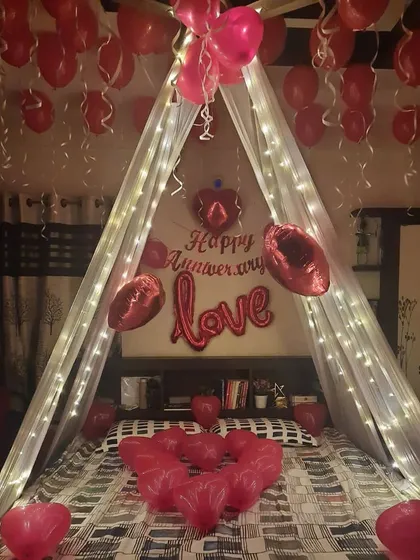A classic bedroom canopy decoration for an anniversary, featuring a cascade of red heart-shaped balloons, fairy lights, and 'LOVE' foil letters above the bed.