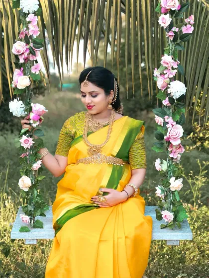 An outdoor traditional shoot featuring a mother-to-be in a bright yellow saree on a floral swing. The natural light and lush greenery make this a vibrant and joyful portrait.