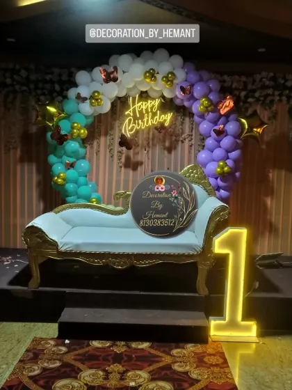 A butterfly-themed stage setup for a first birthday. It features a circular balloon arch in rainbow pastel colors, a "Happy Birthday" neon sign, and an elegant chaise lounge for the little one to sit on for photos.