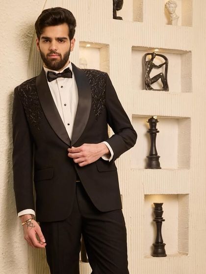 A full-body shot that emphasizes the lean, tailored fit of the classic black tuxedo. The hand-beaded shoulders add a touch of bespoke luxury to this timeless ensemble.