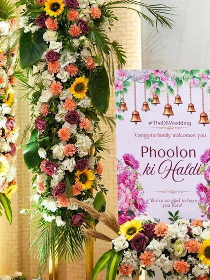 The welcome signage for the 'Phoolon ki Haldi', featuring bright floral graphics and traditional temple bells. This set a festive and welcoming tone for the event right from the entrance.