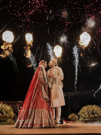 A fairytale moment under the fireworks. The bride's magnificent red wedding lehenga and the groom's classic sherwani create a picture-perfect memory against the night sky.