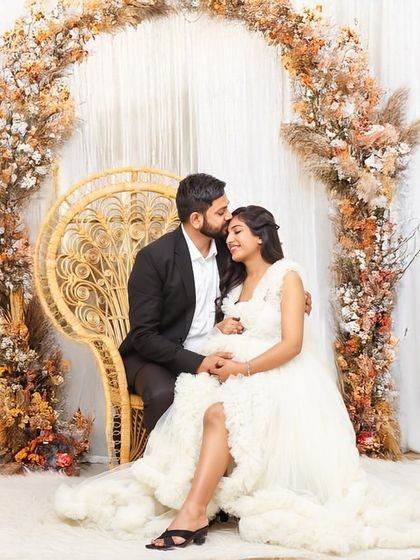 A loving partner kisses the mom-to-be's head in this beautiful studio portrait. They are seated on a wicker chair surrounded by an arch of autumn-colored pampas grass, creating a warm, rustic feel.