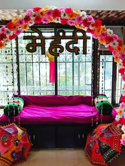 For this home Mehndi function, I created a beautiful and vibrant seating area right by the window. A bright pink sofa is framed by a colorful floral arch and the word "Mehndi" in Hindi script. The setup is completed with traditional Rajasthani umbrellas, making it a perfect photo spot.