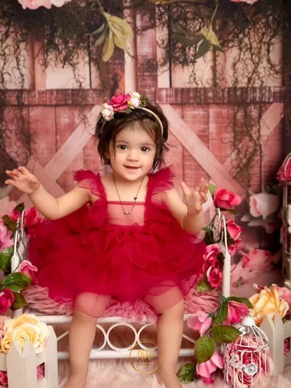 A toddler girl in a festive red dress sits on a miniature bed surrounded by flowers for her birthday shoot.