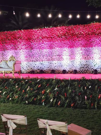 An outdoor reception setting under a canopy of fairy lights. The stage is bathed in a warm golden glow, and the guest tables are decorated with pink accents.