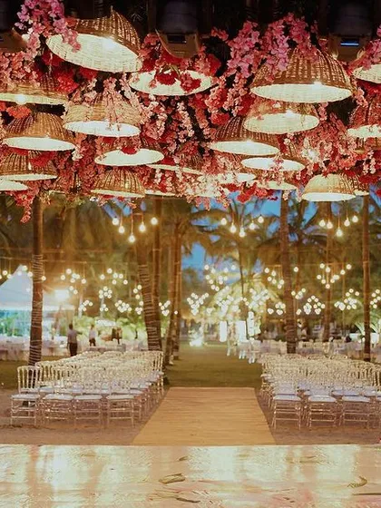 Beach weddings are a whole VIBE. ❤️ For this evening ceremony, we created a canopy of woven basket lamps and pink blossoms, casting a warm, romantic glow over the aisle as dusk settled in.