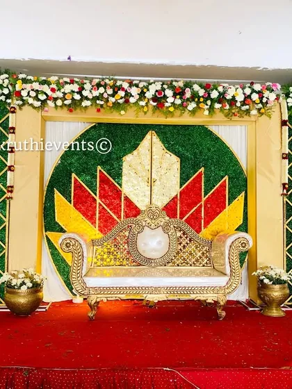 A full stage setup with a central lotus flower backdrop made from marigolds. The stage is flanked by green lattice panels and decorated with brass pots filled with flowers.