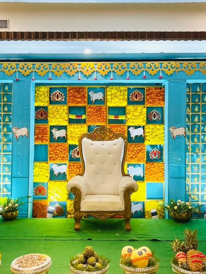 A bright and colorful Seemantham stage with a blue and yellow theme. The backdrop is decorated with cow and lotus motifs, and the foreground is filled with a lavish display of offerings.