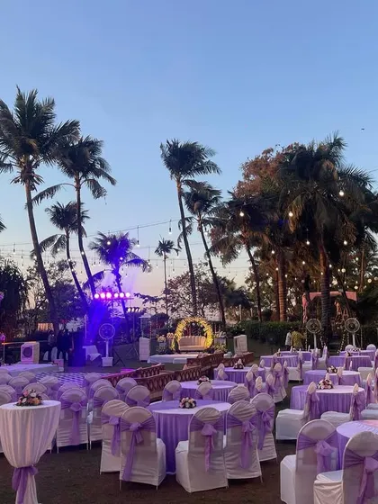 A wide shot of a purple-themed reception setup at twilight. The combination of the open lawn, palm trees, and beautiful lighting creates a stunning ambiance for an evening celebration.