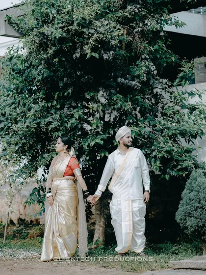 A couple stands hand-in-hand in traditional Brahmin wedding attire. The groom wears a veshti and the bride a classic silk saree.