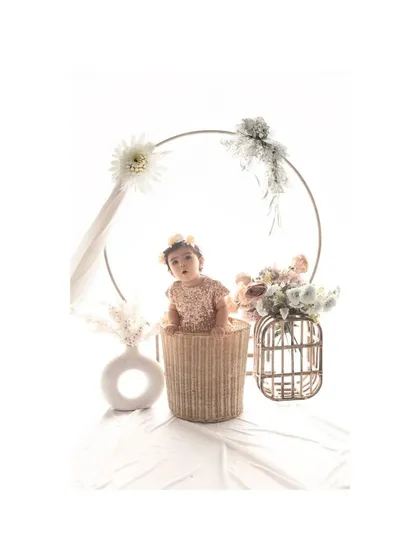 A baby girl sits in a wicker basket, framed by a large golden ring adorned with flowers. This elegant and artistic studio setup creates a beautiful and memorable portrait for a special milestone.