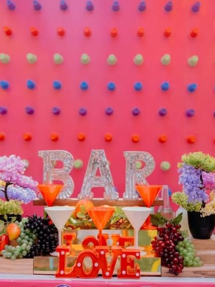 A detailed view of the bar counter. The setup includes fun props like light-up "BAR" and "LOVE" signs, wooden barrels, and fresh fruit, adding to the celebratory vibe.
