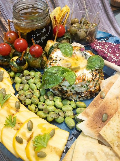 A healthy and vibrant platter with a large herbed cheese ball, edamame, fresh vegetables, and pita bread.