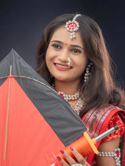 A beautiful portrait of a woman holding a kite and firki. The black background makes her red and black saree and jewelry stand out.
