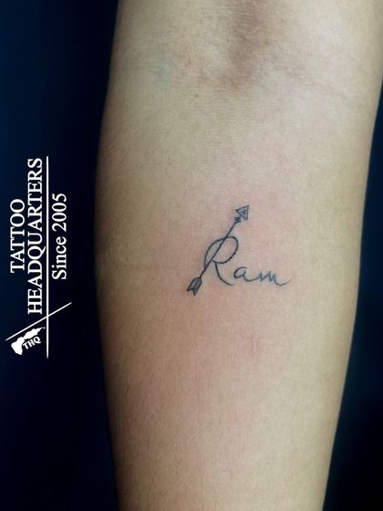 A minimalist tattoo of the name "Ram" in script, with the 'R' cleverly designed as an arrow.