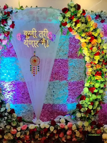 A colorful Dohale Jevan decoration with a checkered flower wall backdrop. The setup includes a floral semi-arch and a sheer cone with a 'Kunitari Yenar Ga!' sign and dreamcatcher.