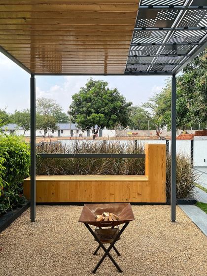 The cozy fireplace and seating area at 'Tranquil Mango Haven'. The pergola features a patterned roof that casts interesting shadows, while the wooden bench and gravel floor create a rustic, comfortable outdoor room.