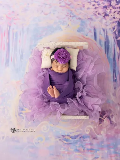 A lavender dream. This newborn is nestled in a bed of purple tulle against a magical, painted backdrop, creating a truly enchanting and artistic portrait.