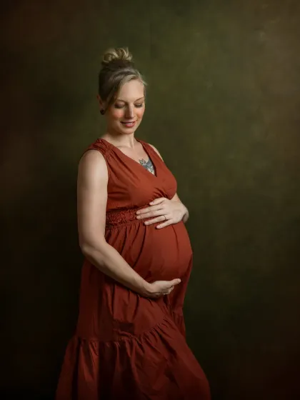 An expecting mother in a rust-colored gown, cradling her belly. The studio lighting creates a beautiful Rembrandt-style effect, with a soft glow illuminating her profile against a dark, painterly background.