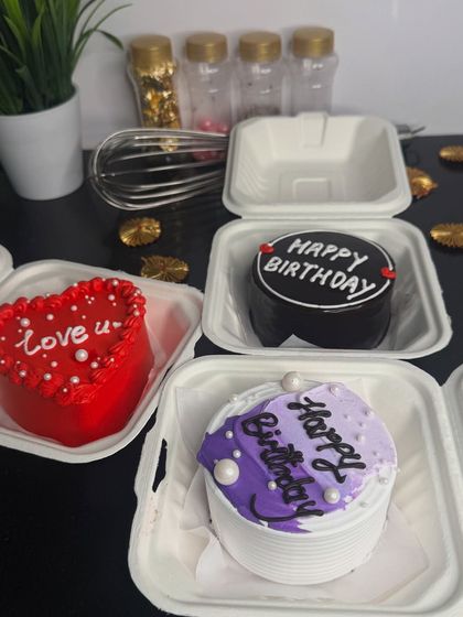 A collection of three different bento cakes, including a heart-shaped "love u" cake, a purple birthday cake, and a chocolate birthday cake. This shows the variety of designs available for these mini treats.