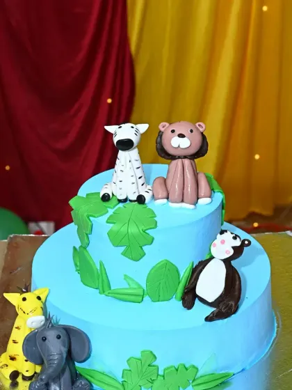 A two-tier jungle animal-themed cake, featuring adorable fondant figures of a lion, zebra, and elephant.