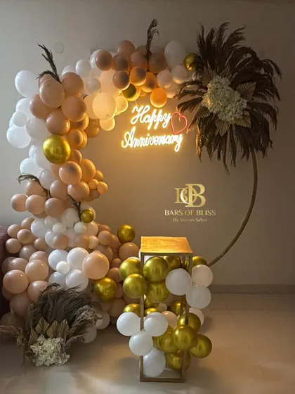 A romantic anniversary setup perfect for a surprise at home. The decor features a half-ring balloon arrangement in soft, neutral tones, accented with florals and a 'Happy Anniversary' neon sign.