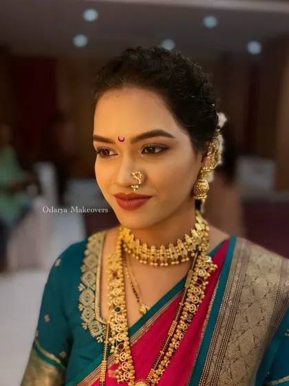 A flawless wedding look for a Maharashtrian bride. The makeup is soft and neutral, allowing the bright colors of her saree and the heavy gold jewellery to stand out.