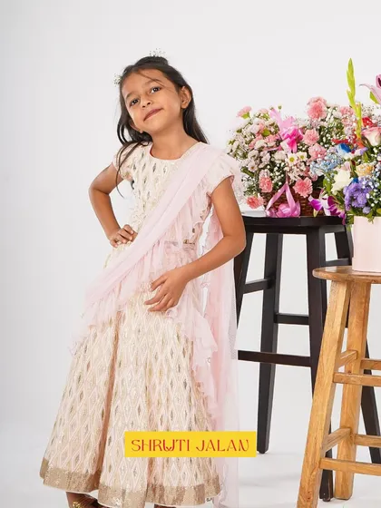 A little princess posing in a cream and gold printed lehenga with a soft pink ruffled dupatta. It's a dreamy and elegant look for a special occasion.