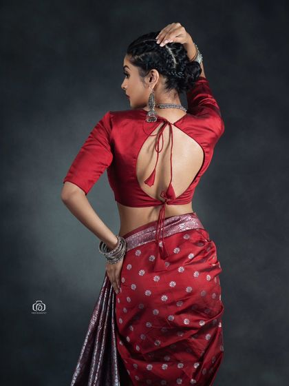 A back-focused shot showing the details of the red blouse and the modern drape of the saree. The lighting highlights the texture of the fabric and the model's form.