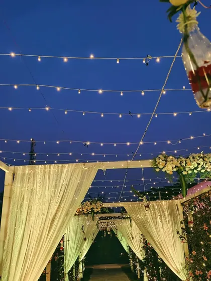A simple yet beautiful pathway for a reception, with strings of fairy lights overhead and elegant table centerpieces featuring lanterns and fresh flowers.