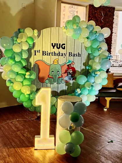 A cute Dumbo-themed first birthday bash for "Yug". This heart-shaped balloon arch in shades of green and yellow frames a custom backdrop, complemented by a large light-up number "1".