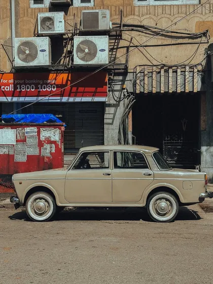 A beautifully maintained, cream-colored Fiat parked against a typical Mumbai backdrop of shops and tangled wires. A slice of real city life.