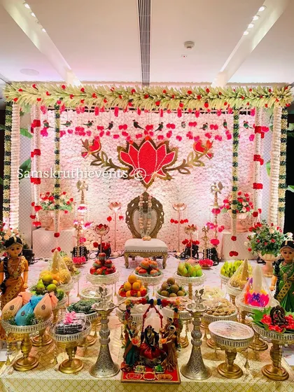 A grand and festive setup for a half-saree or puberty ceremony. The backdrop features a large lotus motif made of flowers, surrounded by traditional brass lamps and a beautiful display of offerings, marking the milestone in style.