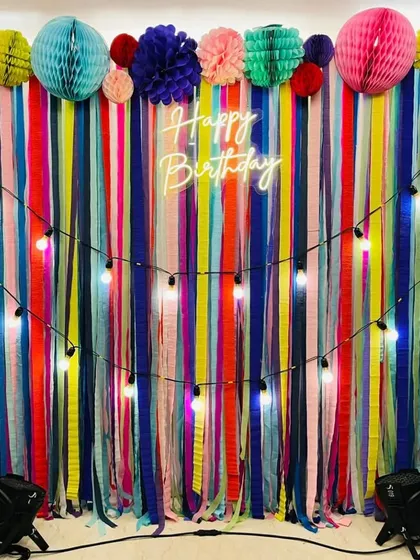 A vibrant and colorful birthday backdrop made of multi-colored ribbons. The setup is enhanced with paper lanterns and string lights for a festive, bohemian feel.
