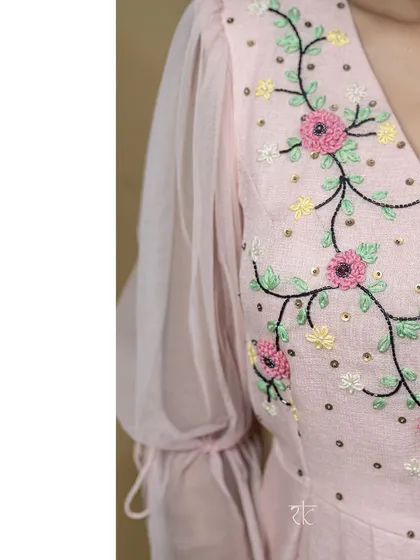 A close-up of a blush pink floor-length dress, showcasing the delicate floral thread embroidery on the bodice. The sheer puff sleeves add a touch of romantic charm.