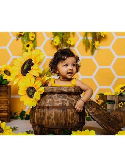 A happy baby girl smiles while peeking out from a wooden barrel filled with sunflowers. The warm yellow tones create a bright and cheerful atmosphere.