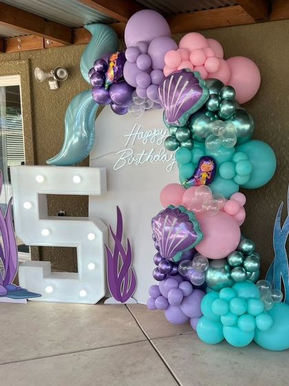 The full view of the mermaid-themed 5th birthday decor, showing the white backdrop, neon sign, and seaweed cutouts.