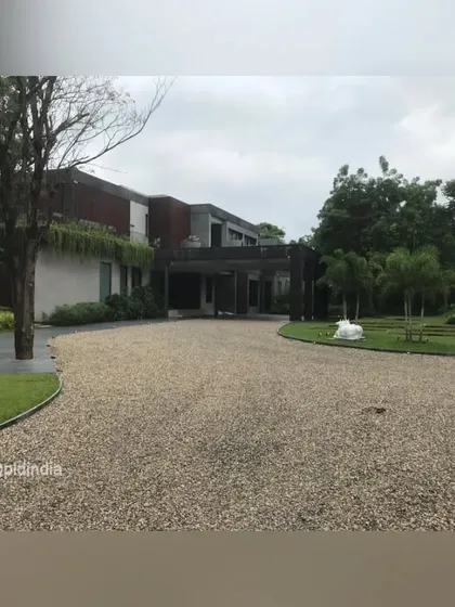 The sweeping gravel driveway leads to the entrance of a modern home in Ahmedabad. The vastness of the entrance court is balanced by the mature trees and lawns that frame it.
