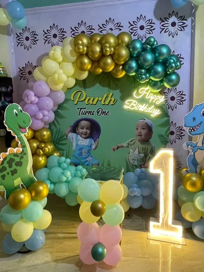 A cute dinosaur-themed first birthday for Parth. This home setup includes a personalized photo backdrop, dinosaur cutouts, a light-up number '1', and a colorful balloon arch with gold accents.