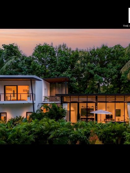 An exterior shot of a modern luxury villa at sunset, nestled in a lush, green environment. The large glass walls and open design blur the lines between indoors and outdoors.