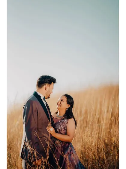 A happy couple in a field of tall, golden grass, smiling at each other. The warm, glowing light gives this pre-wedding photo a dreamy and romantic feel.