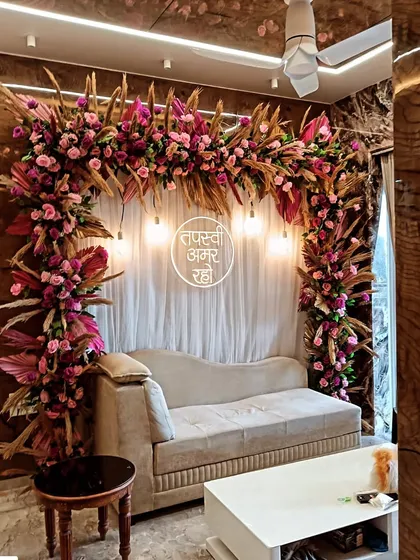 A beautiful floral arch in shades of pink and magenta, designed for a Tapasvi ceremony in a living room. The warm lighting and dried palm leaves create a sophisticated and modern look.