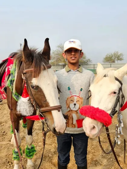A rider in a Christmas sweater poses with two of our horses, who are also dressed up for the occasion with festive gear.