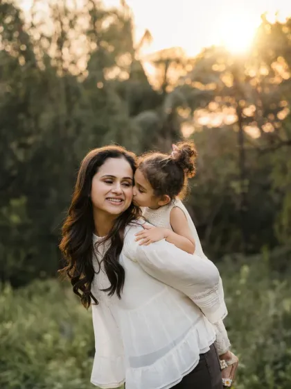 A daughter gives her mom a sweet kiss on the cheek during a piggyback ride. This golden hour photo captures a beautiful mother-daughter bond.