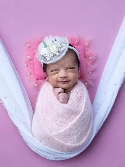 A peaceful, smiling portrait of the baby swaddled in a pink wrap and suspended in a soft white sling.