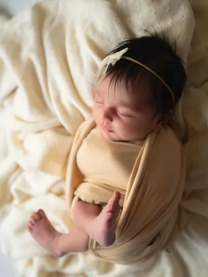 A simple and timeless portrait of a baby wrapped in a soft, butter-yellow swaddle, sleeping on a bed of cream-coloured fabric.